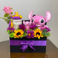 Tagerelsa3ada Angel and Stitch gift box with large pink Angel, mini Stitch, sippy cup, sunflowers, purple daisies, and greenery in a black box with purple ribbon.