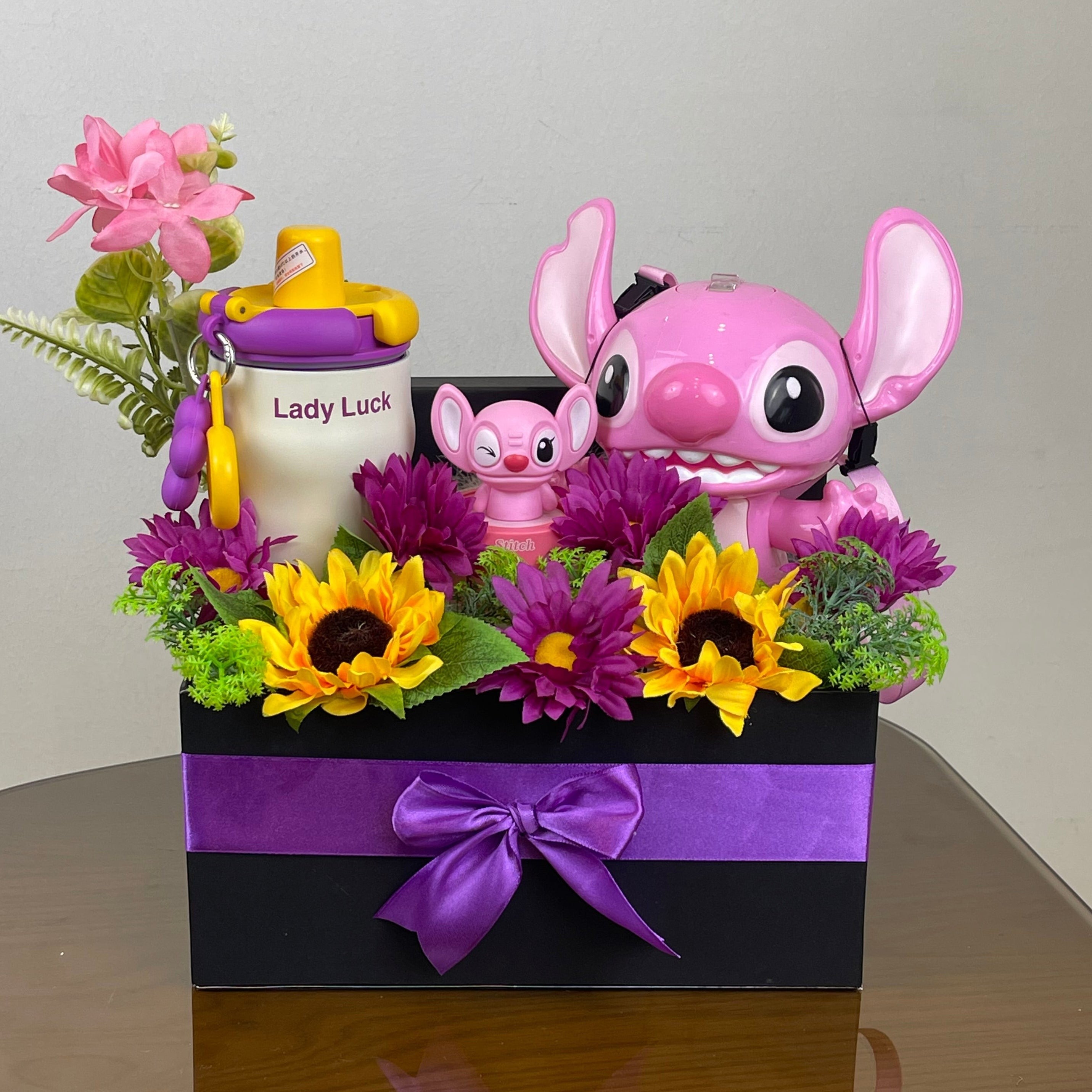 Tagerelsa3ada Angel and Stitch gift box with large pink Angel, mini Stitch, sippy cup, sunflowers, purple daisies, and greenery in a black box with purple ribbon.