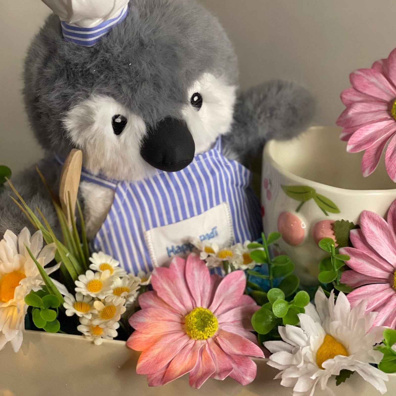 Tagerelsa3ada Chef Penguin plush toy wearing a chef hat and apron, surrounded by pink and white daisies and greenery.