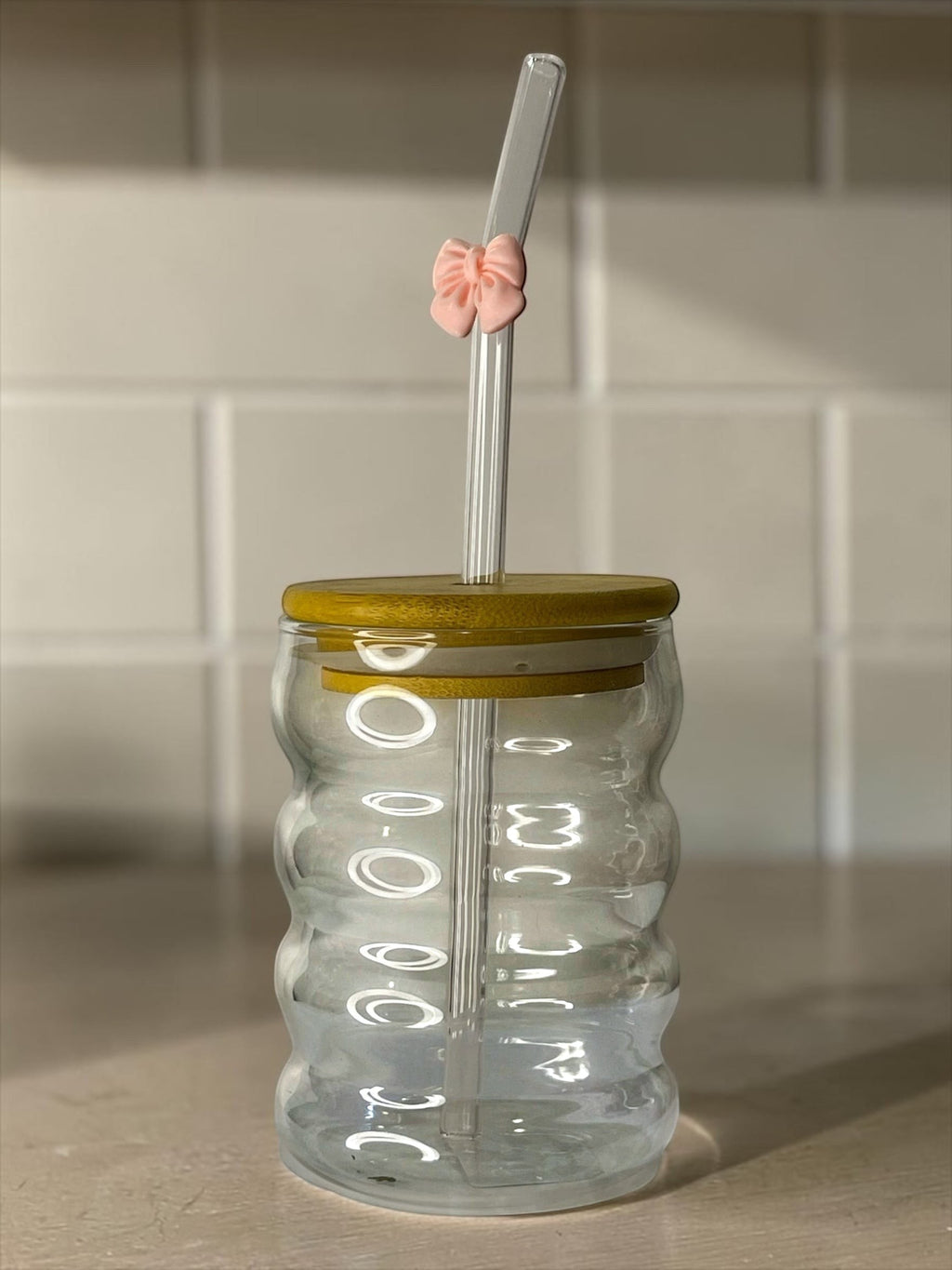 Tagerelsa3ada clear glass tumbler with a ribbed design, bamboo lid, and a clear straw adorned with a small pink bow.