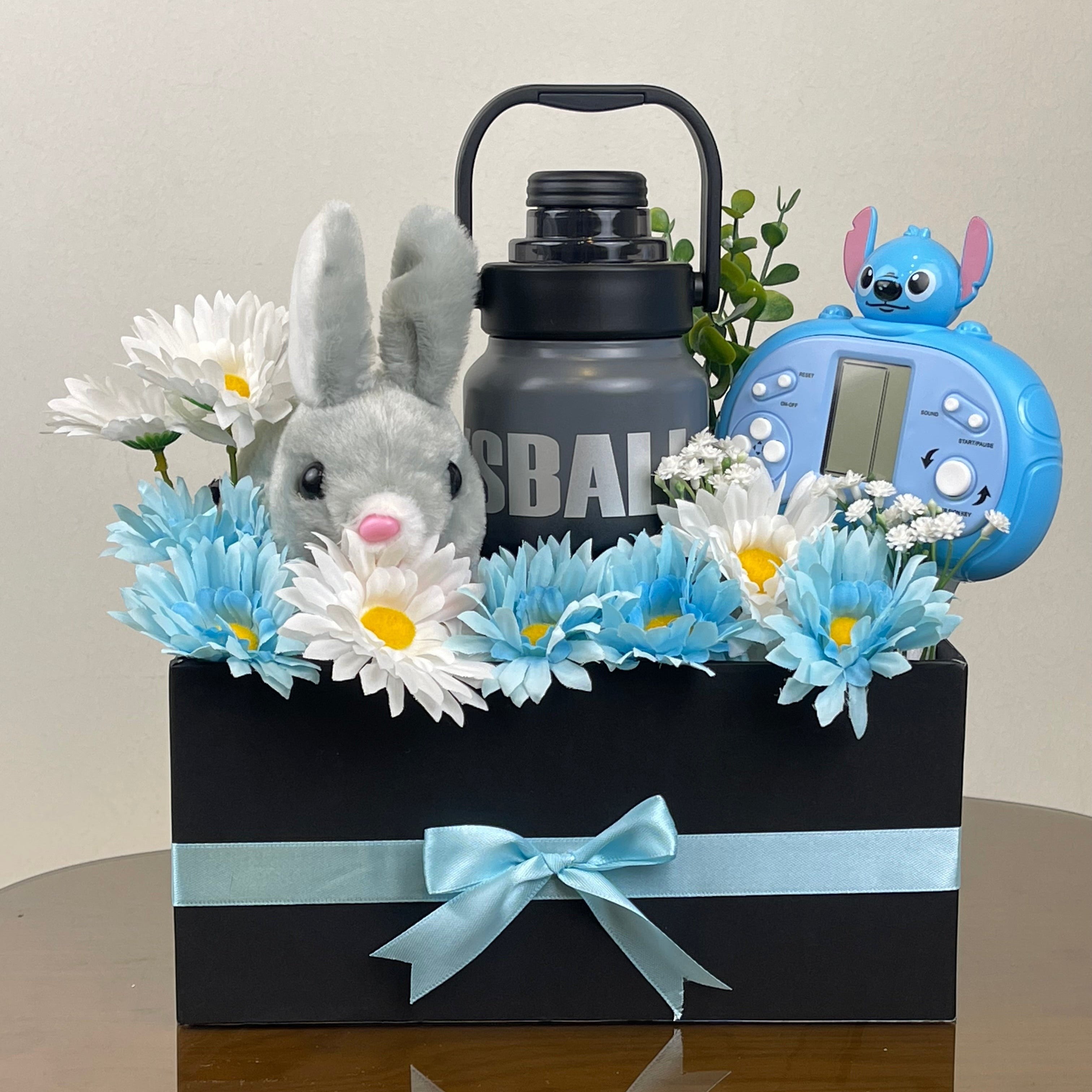 Tagerelsa3ada gift basket with plush bunny, blue & white daisies, grey water bottle, and blue Stitch alarm clock, tied with a light blue ribbon.