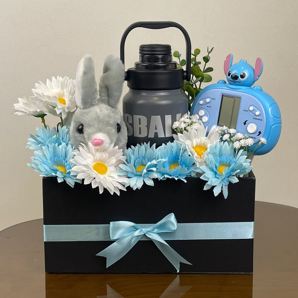 Tagerelsa3ada grey plush bunny, blue and white daisies, grey water bottle, and blue toy in a black gift box with a light blue ribbon.