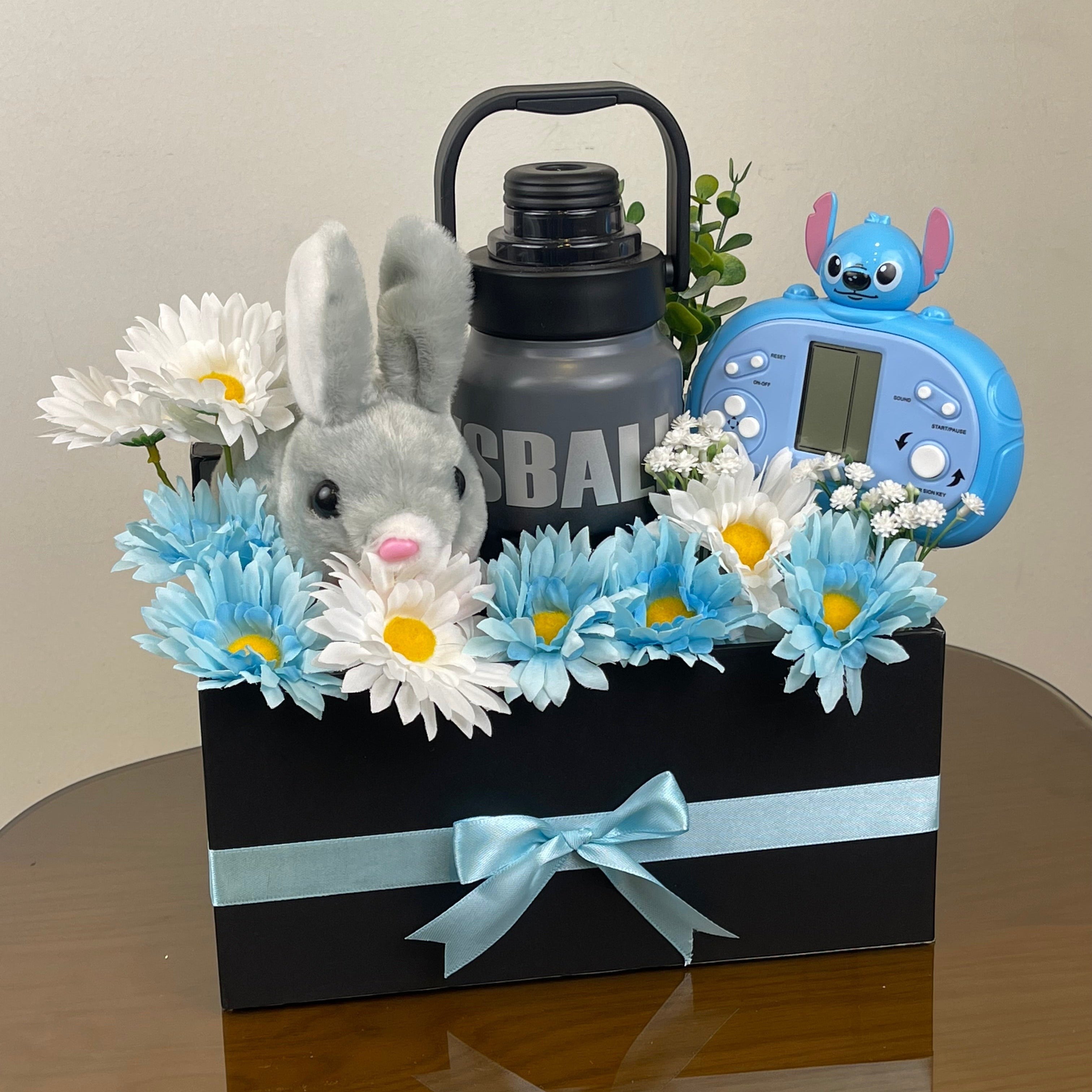 Tagerelsa3ada grey plush bunny, blue and white daisies, grey water bottle, and blue toy in a black gift box with a light blue ribbon.