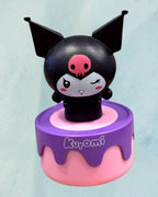 Tagerelsa3ada Kuromi figurine with black ears, pink skull nose, winking eye, on a pink and purple cake-shaped base.