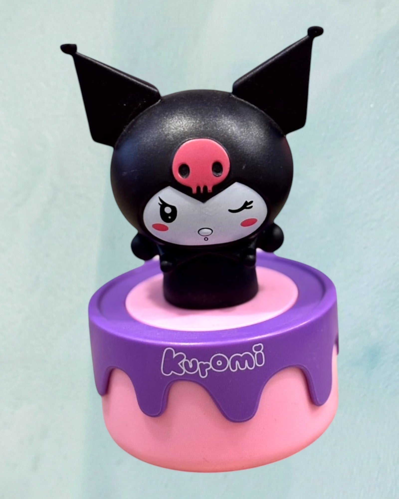 Tagerelsa3ada Kuromi figurine with black ears, pink skull nose, winking eye, on a pink and purple cake-shaped base.