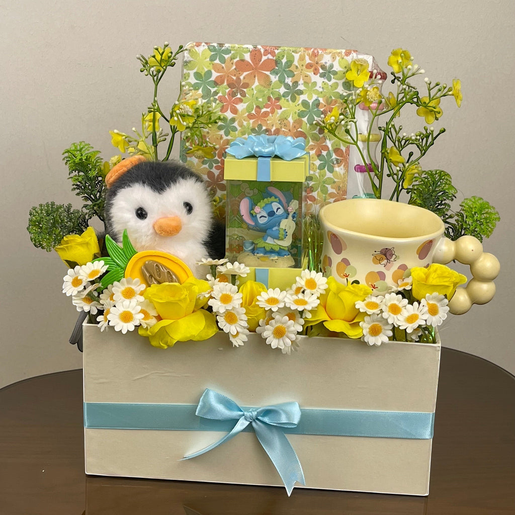 Tagerelsa3ada Penguin and Stitch gift box with plush penguin, Stitch figurine, mug, yellow roses, daisies, and decorative foliage in a cream box with blue ribbon.