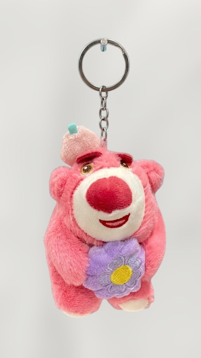 Tagerelsa3ada pink plush bear keychain holding a purple flower, with a small blue square on its head.