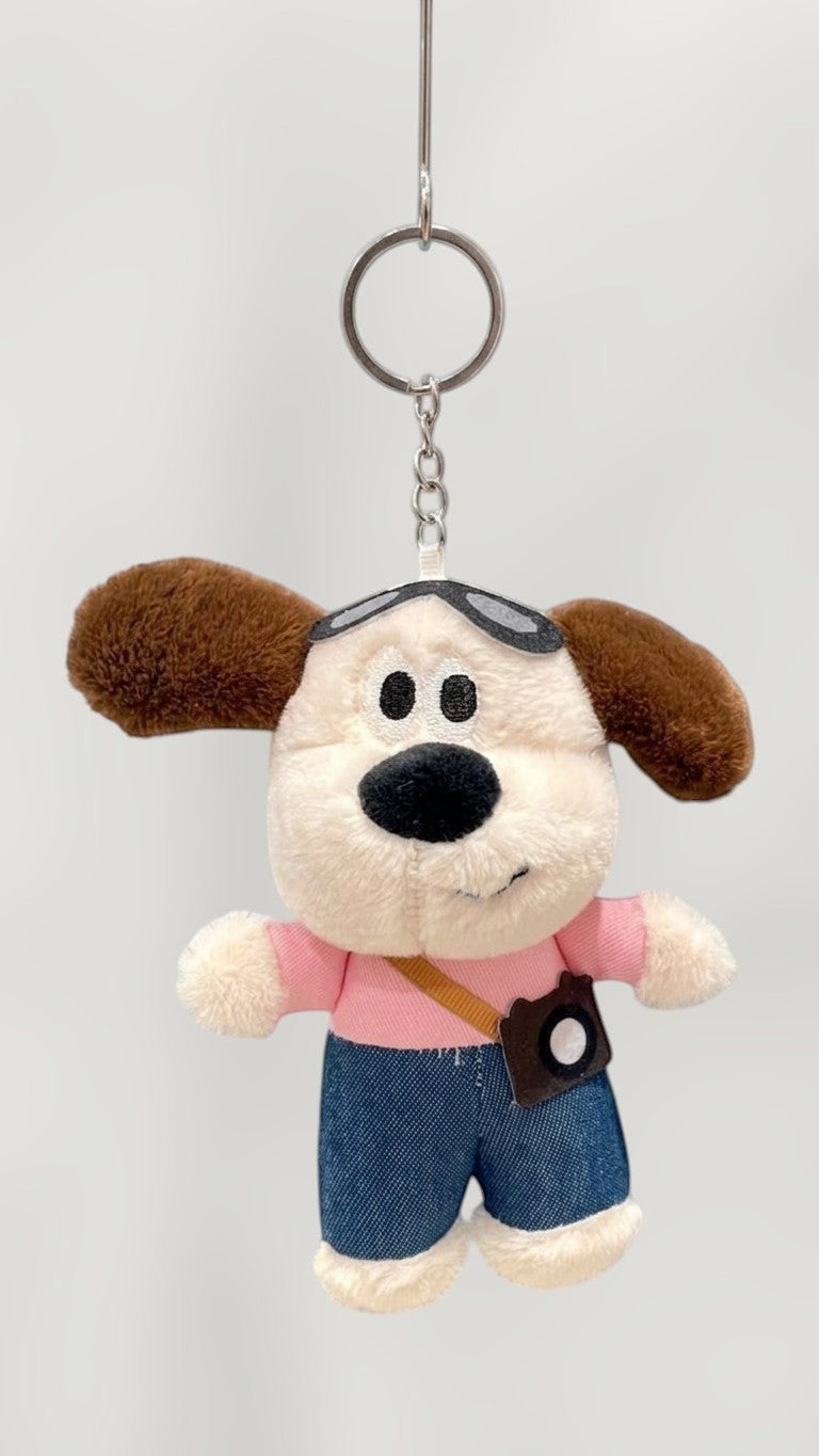 Tagerelsa3ada plush dog keychain with aviator goggles and camera accessory, wearing pink shirt and denim shorts.
