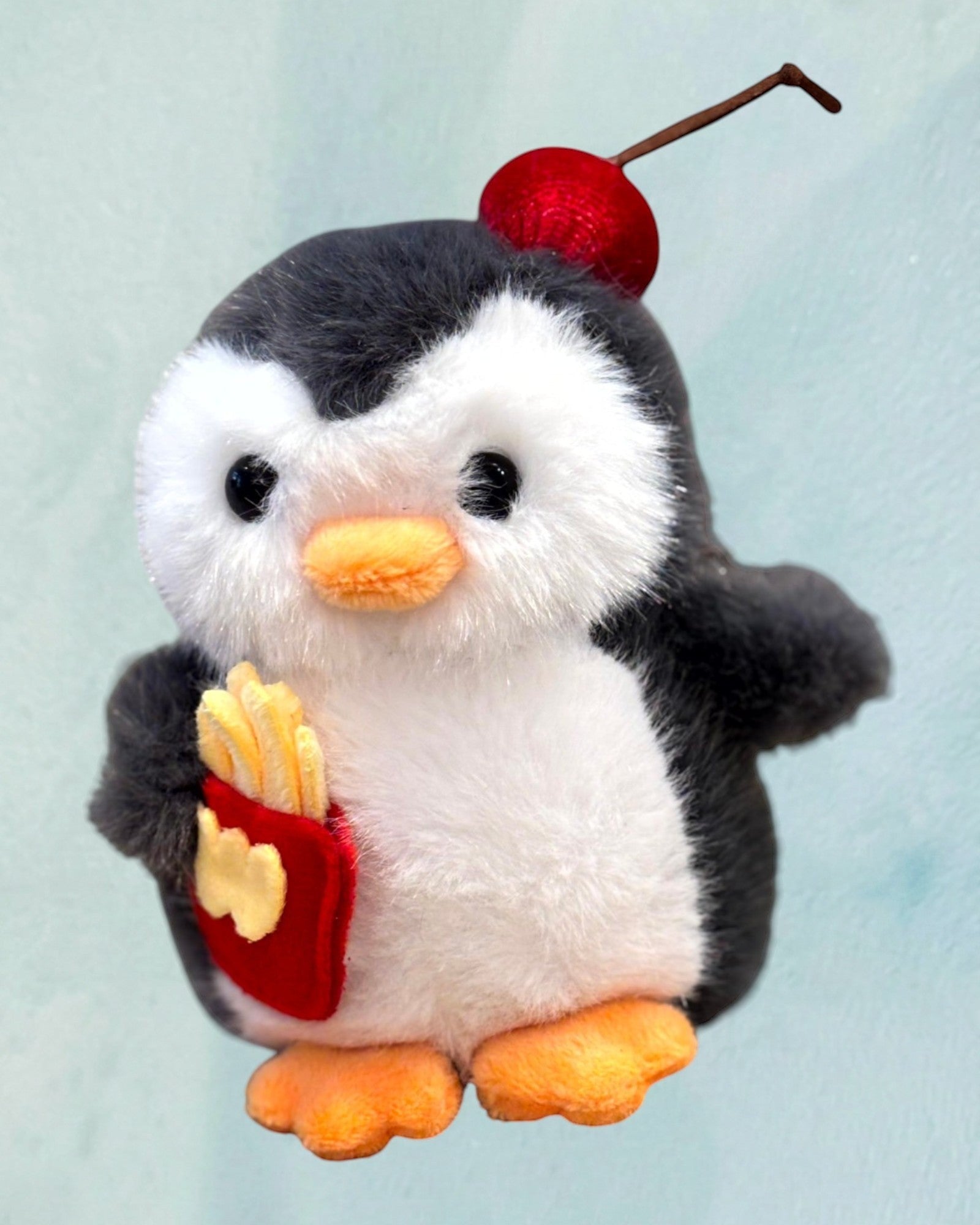 Tagerelsa3ada plush penguin toy with fuzzy white belly, holding french fries, topped with a cherry.