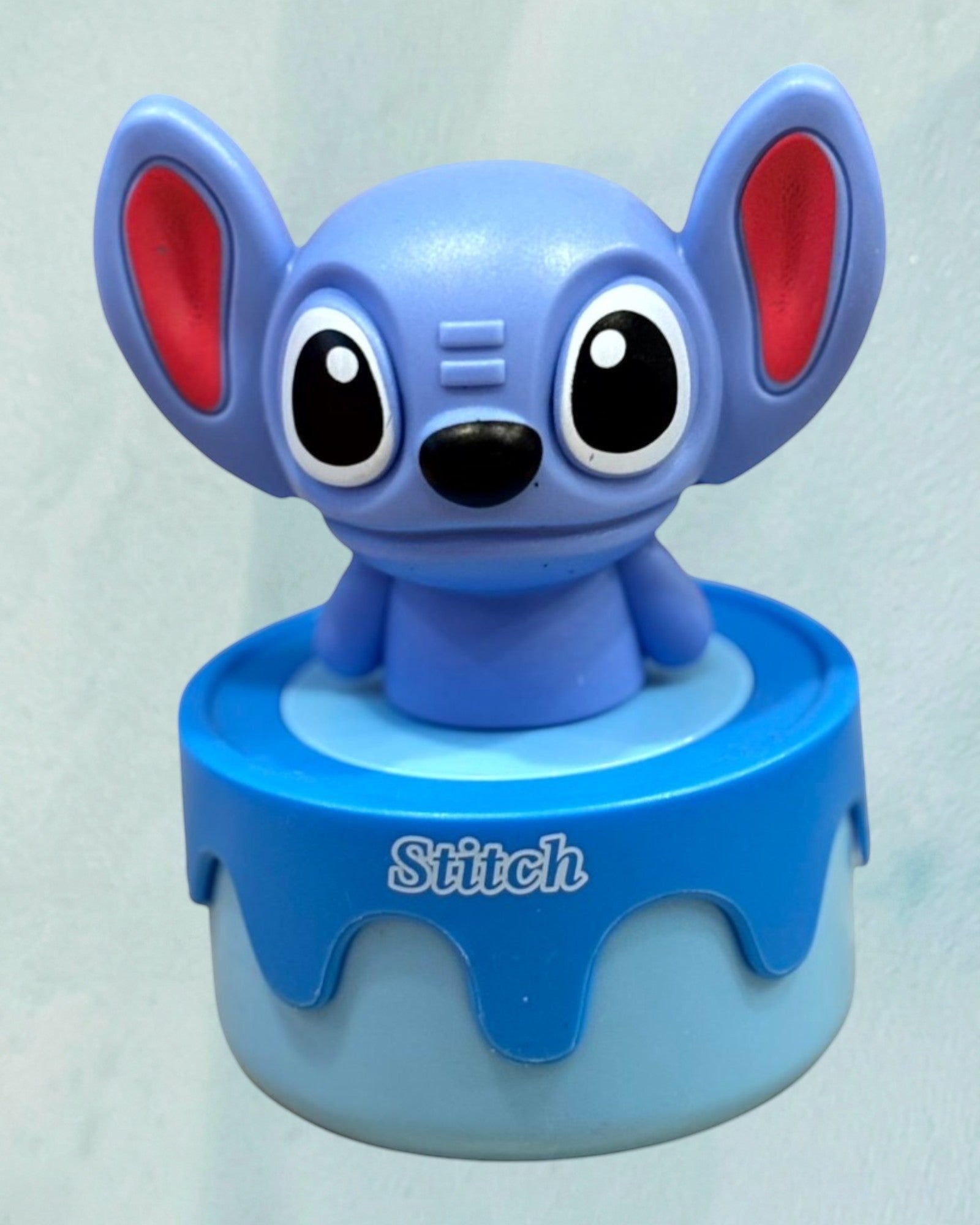 Tagerelsa3ada Stitch figurine, a small blue character with large ears and red inner ears, on a blue base with the name Stitch.