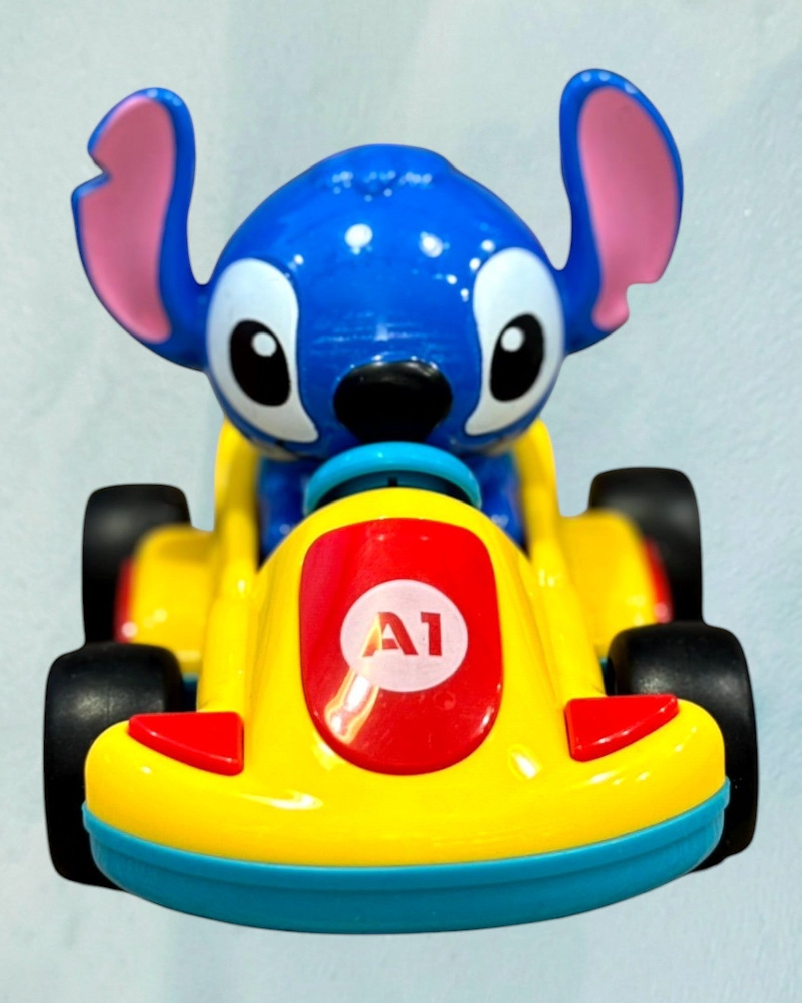 Tagerelsa3ada blue Stitch character in a yellow plastic racing car toy with black wheels and red accents.