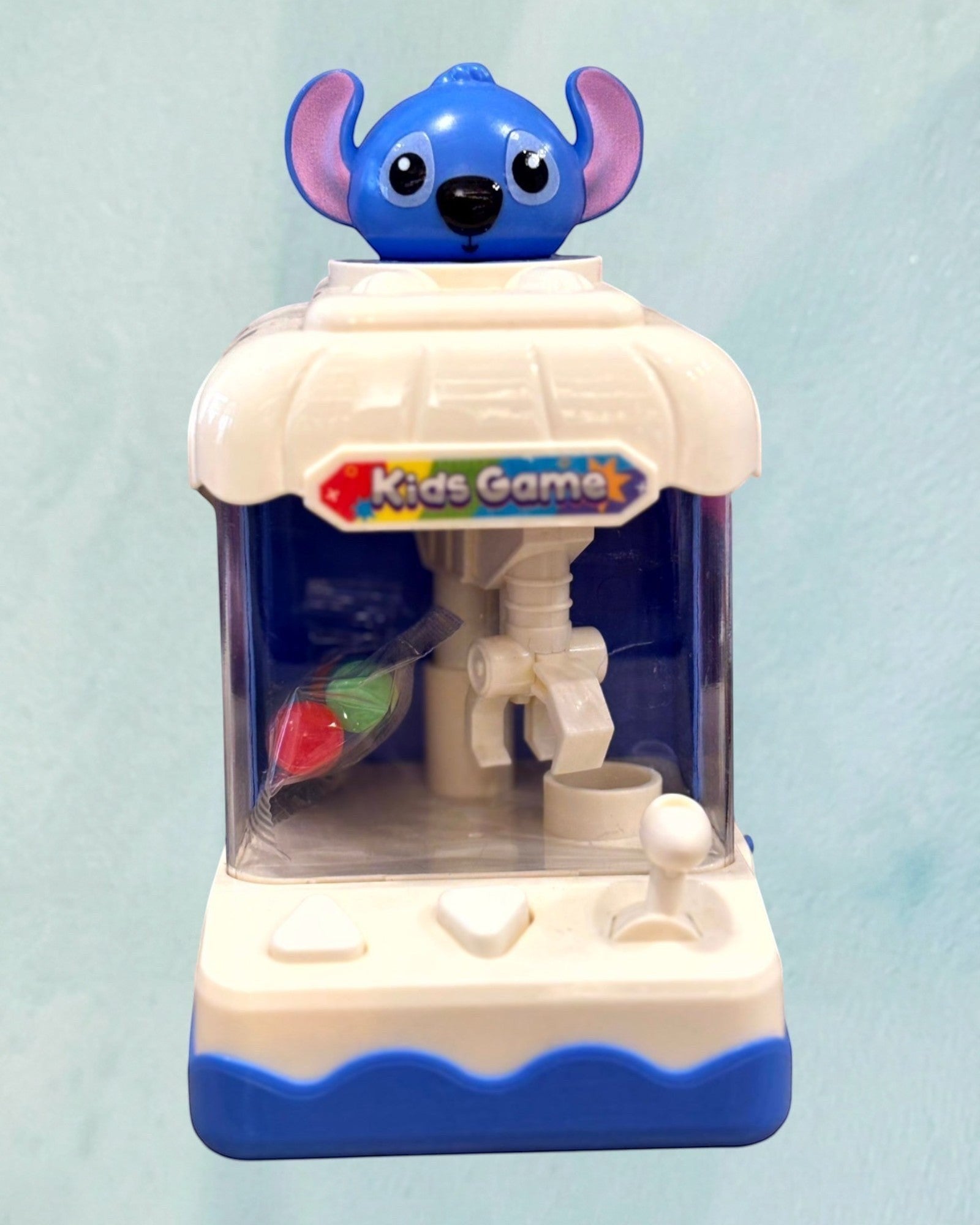 Tagerelsa3adaHere blue and white mini kids game claw machine toy with a blue character on top and colorful prizes inside.